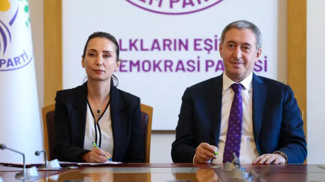 Yan yana duran bir kadın ve erkek. Arkada DEM Parti logosu var