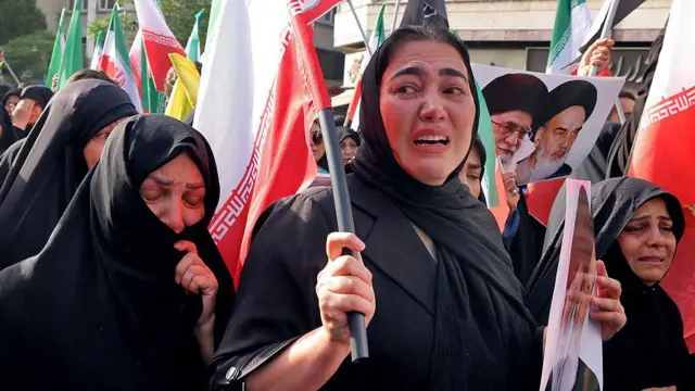 İran'da siyah çarşaf içinde kadınlardan üçü ağlıyor, ellerinde fotoğraflar ve bayraklar var.