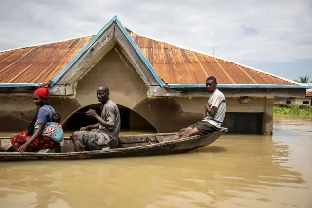 Deux hommes et une femme portant un bébé dans le dos dans une pirogue avec en arrière plan un bâtiment