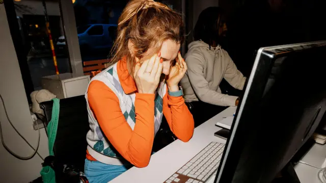 Mulher de blusa laranja de mangas compridas e colete cinza e branco na sua mesa de trabalho, com a cabeça entre as mãos