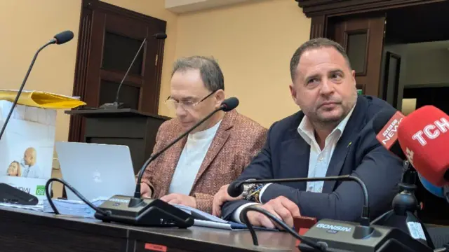 У залі засідань за столом із мікрофонами сидять двоє учасників. Ліворуч — адвокат Ігор Фомін, працює з ноутбуком і документами. Праворуч — Андрій Єрмак, сидить із руками на столі, перед ним мікрофони медіа. На столі лежать папери та мікрофони.