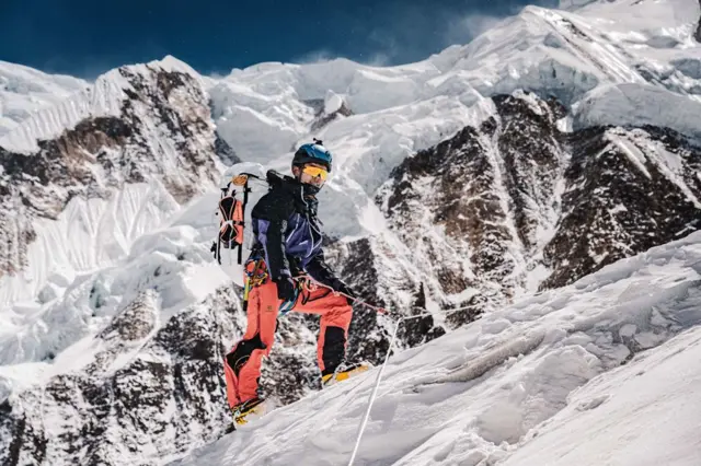 Nima, in climbing gear wearing a large backpack, scales a cliff with a rope