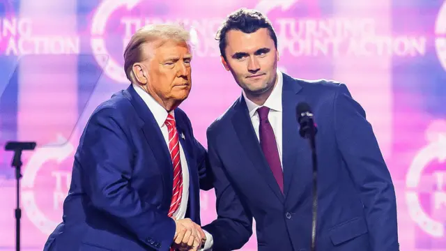 Charlie Kirk (à droite) sur scène avec le président américain Donald Trump lors de l'America Fest 2024 à Phoenix, en Arizona, le 22 décembre 2024. Les deux hommes se tiennent par la main, tandis que Trump a posé sa main libre sur le dos de Kirk. Tous deux sont penchés l'un vers l'autre et portent des costumes bleus. L'arrière-plan est rose et violet, avec l'inscription « Turning Point Action » sur l'écran.
