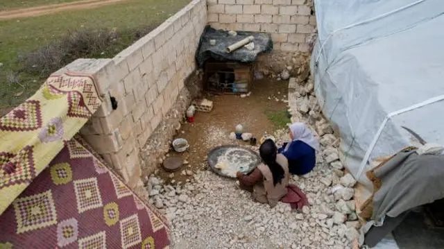 تصویر یک زوج سوری که در میان ویرانه‌های خانه شان در کنار خیمه نشسته‌اند. این زوج در حال صرف غذا در روی زمین هستند. 