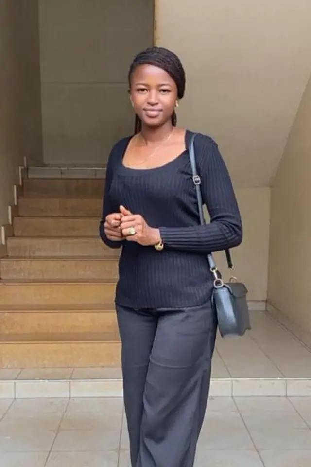 Noella stands in front of a set of stairs, in a black top and trousers, carrying a black bag. Her hair has been tied up, and she smiles at the camera. 