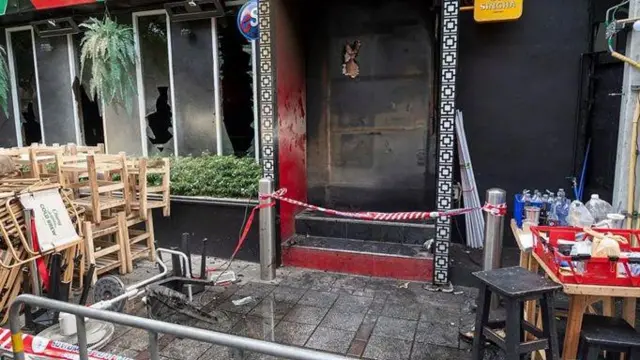 Вхід до бару після пожежі. Територію перед входом огороджено сигнальною стрічкою. Видно пошкоджені скляні вітрини, закопчені стіни та сліди впливу вогню на фасаді. Біля входу розміщені меблі та господарські предмети.