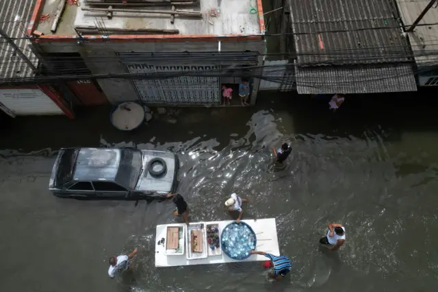 Foto aérea mostra rua inundada e pessoas transportando água e mantimentos em tábua