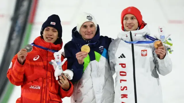 3人がジャンプ台を背に横並びで立ち、笑顔でメダルを手にしている。二階堂とトマシャクは大会マスコットのぬいぐるみも持っている