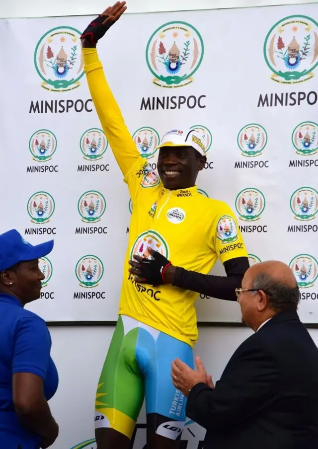 Jean Bosco Nsengimana wambaye umupira w'umuhondo n'ikabutura y'icyatsi kibisi n'ubururu, n'akagofero k'umuhondo yazamuye akaboko