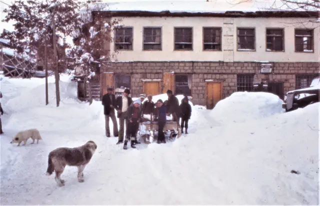 Pülümür'de iki katlı bir ev, önündeki bir grup insan ve iki köpek görülüyor. Etraf ise karlarla kaplanmış.