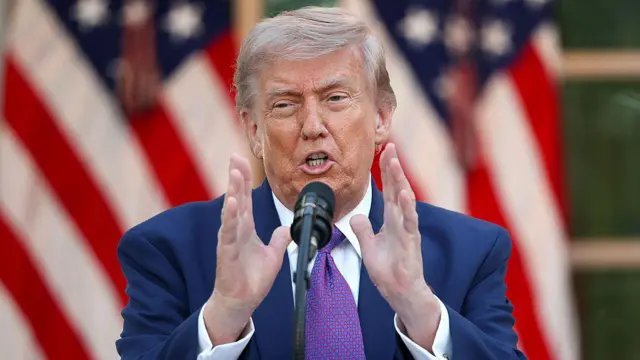 Trump lacivert ceket giyinmiş ve mikrofon önünde iki elini uzatmış halde Amerikan bayrağı önünde konuşuyor