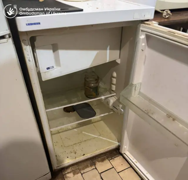 Дуже брудний маленький холодильник, всередині видно бруд, якісь речі. Всі полички майже пусті, на одній стоїть відкрита банка з чимось всередині