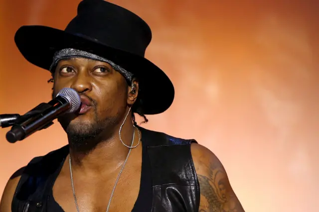 D’Angelo is performing on stage, wearing a wide-brimmed black hat and a black leather vest over a deep V-neck top. A silver chain necklace is visible around the neck, and the left arm shows a detailed tattoo design. He is positioned near a microphone with a black foam cover. The background features a warm orange gradient, creating a dramatic and vibrant concert atmosphere.