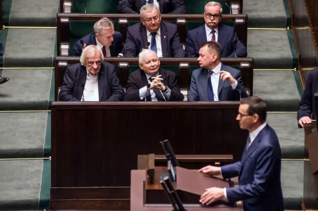 У залі парламенту кілька політиків, в тому числі Ярослав Качинський і тодішній міністр оборони Маріуш Блащак, сидять на зелених лавках і спілкуються між собою під час засідання. На передньому плані видно трибуну, де виступає Моравецький.