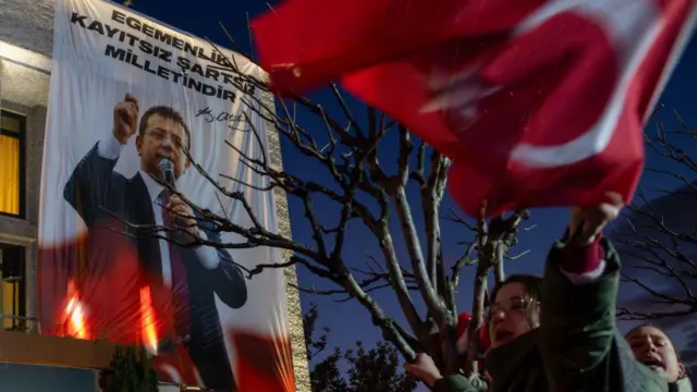 Bir banaya asılmış Ekrem İmamoğlu posteri, önünde Türkiye bayrağı sallayan bir kadın