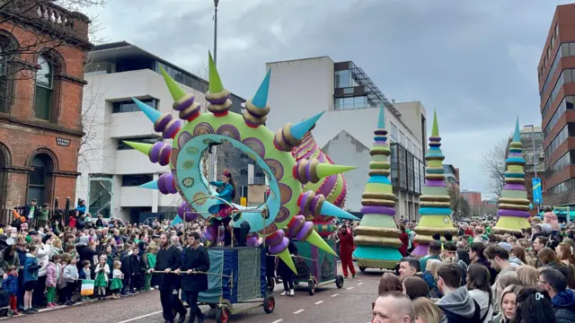 Di image capture a lively parade scene wit a crowded street of spectators. A float featuring a large circular structure wit spikes and intricate patterns take centre stage, accompanied by oda colourful, tiered floats in bright hues. Performers in vibrant costumes stand on di floats, adding to di festive atmosphere. Di crowd dey dressed in celebratory attire, reflecting di excitement and community spirit of di event. For di background, e get a mix of modern and older architecture, wit a visible street sign reading 'VICTORIA STREET' on a brick building to di left
