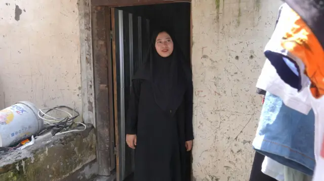 Siti Aminah dengan baju terusan hitam dan jilbab hitam, bersandar ke pintu.