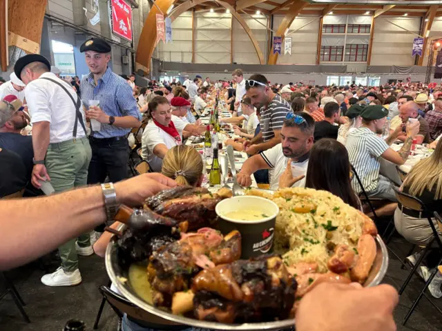Em primeiro plano, vê-se um grande prato de carne de porco e batatas. Ele é servido em uma bandeja de metal por um homem com um relógio de pulso. Ao fundo, centenas de pessoas sentam-se em longas mesas, com garrafas de vinho dispostas ao longo do centro das mesas.