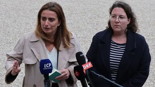 白い壁の前に立った女性2人が記者たちからマイクを向けられている。左側の女性はベージュのコートを着て、左手に緑色の手帳を持って話している。右側の女性は紺色のジャケットを聞いて、左側の女性の話を聞いている