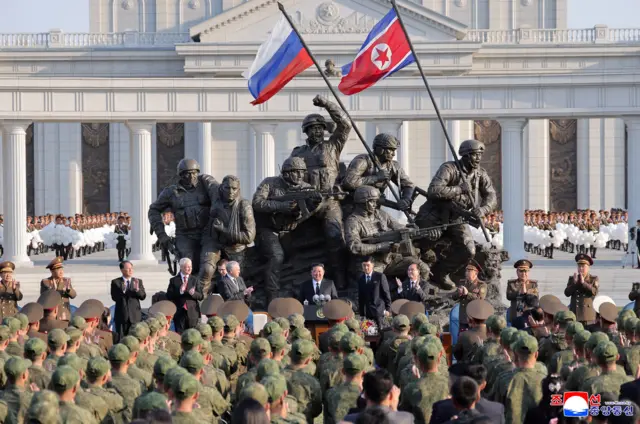 Военный мемориал с флагами РФ и КНДР, перед ним стоят официальные лица и военные.
