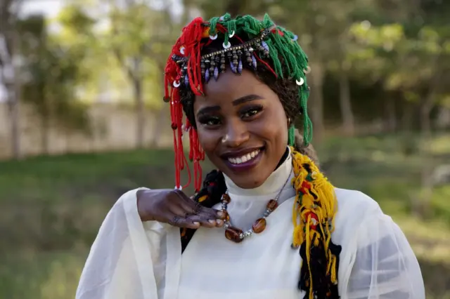 A woman wearing a tasselled red, green and yellow headdress smiles.