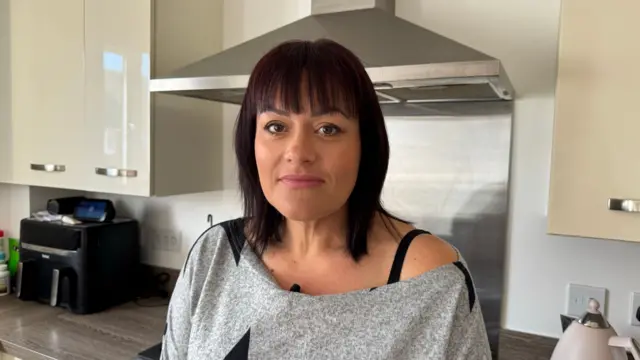 Hayley Dawe vestindo camiseta cinza com sua cozinha ao fundo, ela tem cabelo liso escuro com franja e olhos castanhos