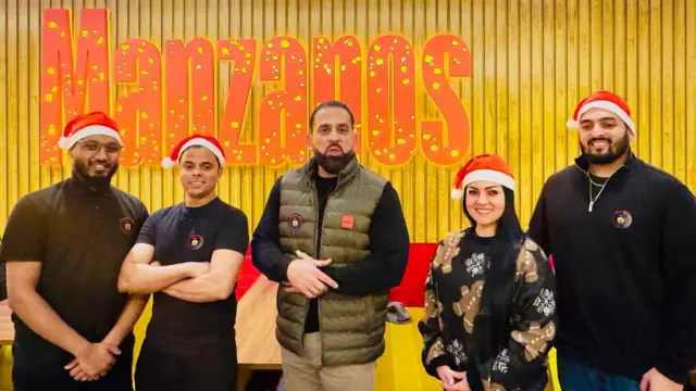 Asam Aziz (centro) es el dueño de Manzanos. Su hijo, Kasim Aziz, está en el extremo derecho. La foto muestra a Asam Aziz rodeado de familiares o empleados con gorros navideños frente a un gran cartel de Manzanos.