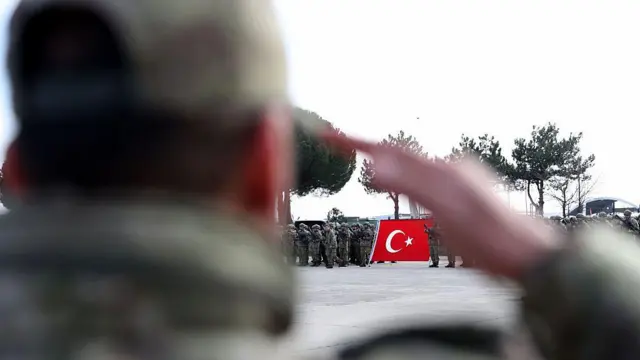 Arkası dönük bir askerin selam yaptığı kolunun arasından Türk bayrağı tutan tabur uzakta görülüyor.