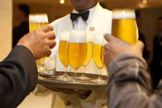 Duas pessoas pegam taças de chope de bandeja de garçom, que usa gravata borboleta