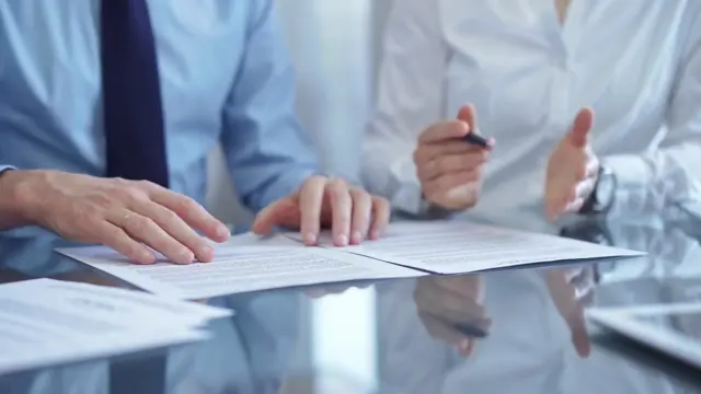 Foto de uma mesa com papeis e duas pessoas com as mãos próximas como se estivessem discutindo sobre alguma documentação.