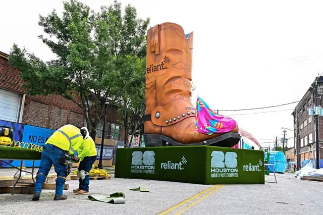 Una calle en Houston, Texas en la que se está instalando una zona para que fanáticos del fútbol puedan disfrutar los partidos. Hay ds trabajadores instalando unas mesas y detrás de ellos se ve una inmensa bota texana de adorno que está instalada en la mitad de la calle