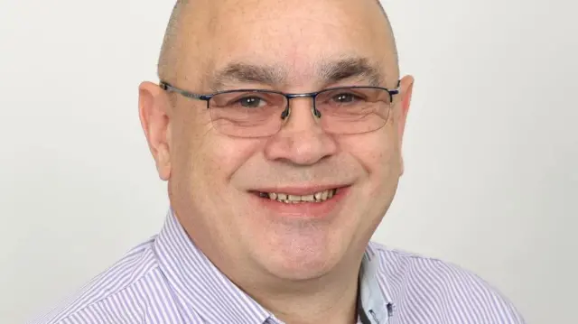 Gary Johannes lleva una camisa a rayas moradas y blancas y gafas, sonriendo a la cámara.