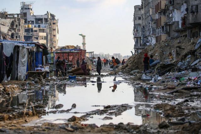 Palestinos desplazados caminan alrededor de un charco frente a edificios y tiendas de campaña destruidos en Jan Yunis, en el sur de la Franja de Gaza, el 16 de mayo de 2024.