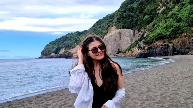 Дівчина на побережжя біля води, ззаду видно скелясту гору, на якій ростуть зелені дерева та інші насадження