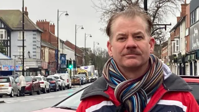 Una foto de Michael Moody, un hombre de mediana edad con cabello castaño y bigote. Lleva una bufanda multicolor y una chaqueta roja, blanca y azul. Al fondo se ve una calle transitada, con una hilera de farolas de aspecto elegante con sus finos postes y lámparas redondas.