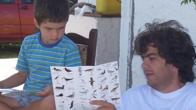 A young boy sitting outdoors and looking at illustrated bird identification sheets, with an older male holding a guide showing various bird species.