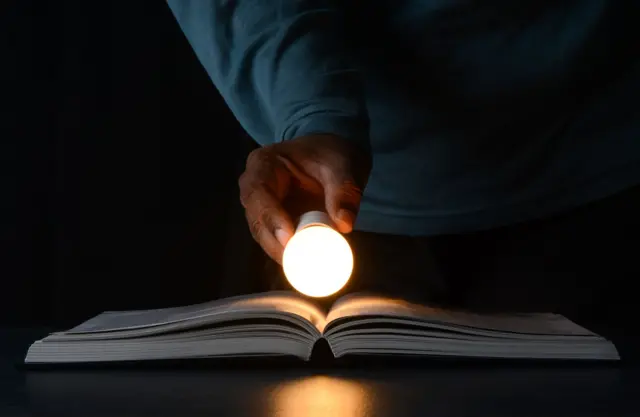 مفهوم الأفكار الإبداعية والتعلم والابتكار. رجل يحمل مصباحاً مضيئاً على كتاب مفتوح. تظهر فقط يد الرجل التي تبدو سمراء كما يبدو أنه يرتدي فميصاً أزرق اللون، مع المصباح والكتاب.