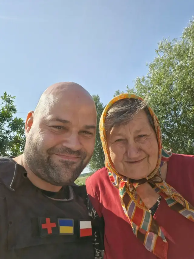 Чоловік у захисному спорядженні фотографується поруч із літньою жінкою в хустці на тлі дерев і ясного неба.