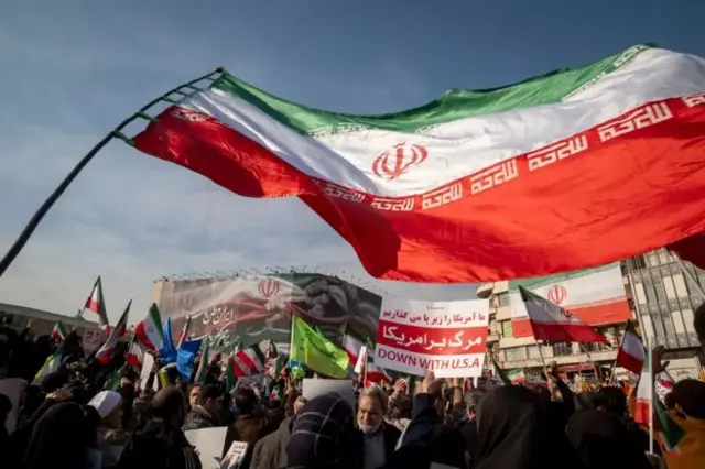 در صحنه‌ای از اعتراضات ایران، پرچم بزرگ جمهوری اسلامی در آسمان به اهتزاز درآمده و شهروندانی با پلاکاردها و پرچم‌هایی در زیر آن دیده می‌شوند. در پس‌زمینه، بنر بزرگی بر دیوار بیرونی ساختمان آویزان است