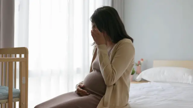 Seorang ibu hamil menangis, menutup wajahnya dengan satu tangan dan tangan lainnya memegang perutnya. Dia duduk di tepi tempat tidur, di depannya ada ranjang bayi terbuat dari kayu.