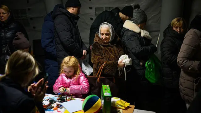 Люди гріються разом в одному з пунктів незламності, січень 2025. На їх фоні виділяється дівчинка в рожевій курточці, яка зайнята малюванням чи конструюванням