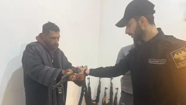 رجل يرتدي معطفاً يسلم سلاحاً بيديه لرجل يرتدي زياً رسمياً مكتوب عليه 