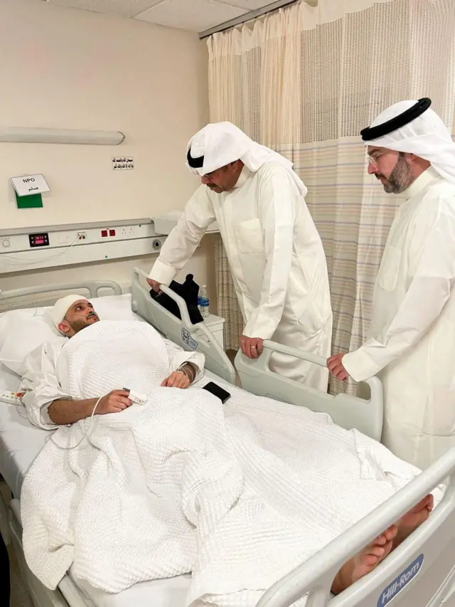 El ministro de Defensa kuwaití, el jeque Abdullah Ali Abdullah al Salem al Sabah, un hombre con túnica blanca mira a una persona en una cama, con la cabeza con vendas.
