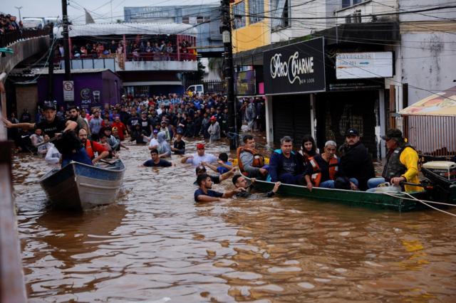 Foto mostra dezenas de pessoas amontadas em uma área seca em frente a uma rua alagada