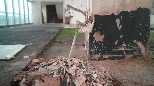 La imagen muestra el interior de un edificio en proceso de demolición o de remodelación a fondo. En primer plano, se amontonan escombros en el suelo: yeso roto, tejas y trozos de hormigón. Cerca se encuentra una columna dañada, con su superficie exterior desportillada, de modo que quedan colgando mallas metálicas y fragmentos de yeso.