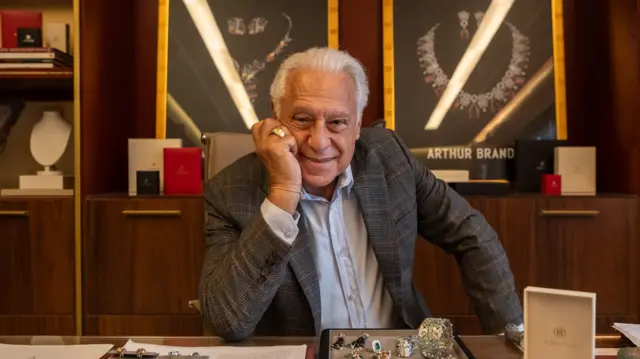 Antonio Fagundes, um homem de cabelos brancos, posa apoiando o rosto na mão atrás de uma mesa com joias e documentos, em um ambiente sofisticado de escritório. Vestindo blazer xadrez e camisa clara, ele sorri para a câmera enquanto, ao fundo, vitrines iluminadas exibem colares de cristais.