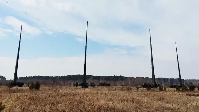 Четыре военных грузовика с высокими антеннами. Вокруг — поле с сухой травой и линия деревьев на заднем плане, над ними — облачное небо.