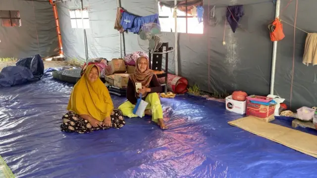 Salamah, memakai kerudung panjang berwarna mustard, dan daster, duduk di atas terpal berwarna biru yang menjadi alas tenda pengungsian. Di sebelahnya juga ada korban lainnya, memakai kerudung cokelat bercorak, baju cokelat, dan celana berwarna hijau terang, memegang handphone di tangan kanannya serta sebuah pouch.