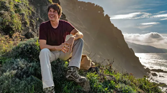 O Prof. Brian Cox está sentado em uma encosta coberta de árvores, com o mar no lado direito da imagem. Ele está vestindo uma camiseta bordô, calça cinza e botas de caminhada. Ele está sorrindo enquanto olha para longe. O Sol está brilhando intensamente ao fundo