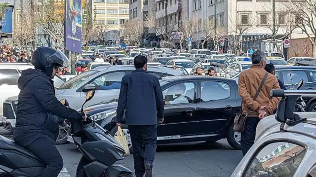 Iranianos tentam atravessar rua em meio ao tráfego intenso em Teerã, capital do Irã, no dia 28 de fevereiro de 2026, após ataques de Israel e EUA. Um homem está em uma scooter e dois outros a pé ao lado de filas de carros.